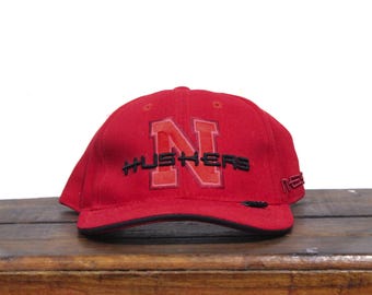 May include: Red baseball cap with a black embroidered "N" and "HUSKERS" in black lettering.