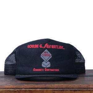 Puede incluir: Gorra de camionero negra con parte trasera de malla y visera plana. La parte delantera muestra texto rojo: "LORNE G. SEIFERT INC." sobre un diamante gris con "EST 1959" y "Concrete Contractors".