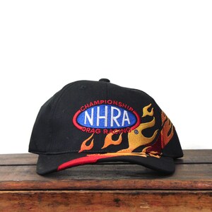 May include: Black baseball cap featuring the text "CHAMPIONSHIP NHRA DRAG RACING" in a blue and red oval design. The cap has a flame design in yellow, orange, and red.