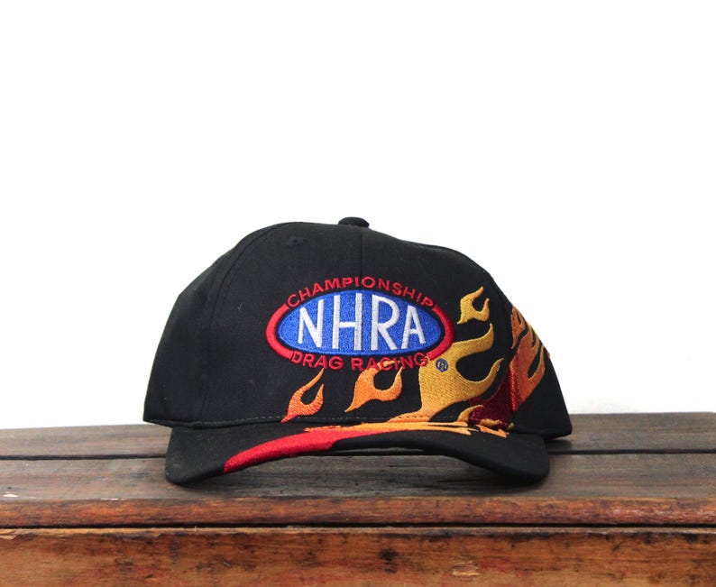 May include: Black baseball cap featuring the text "CHAMPIONSHIP NHRA DRAG RACING" in a blue and red oval design. The cap has a flame design in yellow, orange, and red.