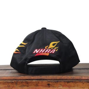 May include: Black baseball cap with embroidered flames in yellow and orange. The cap features the text "NHRA" in red and orange. The cap is sitting on a wooden surface.
