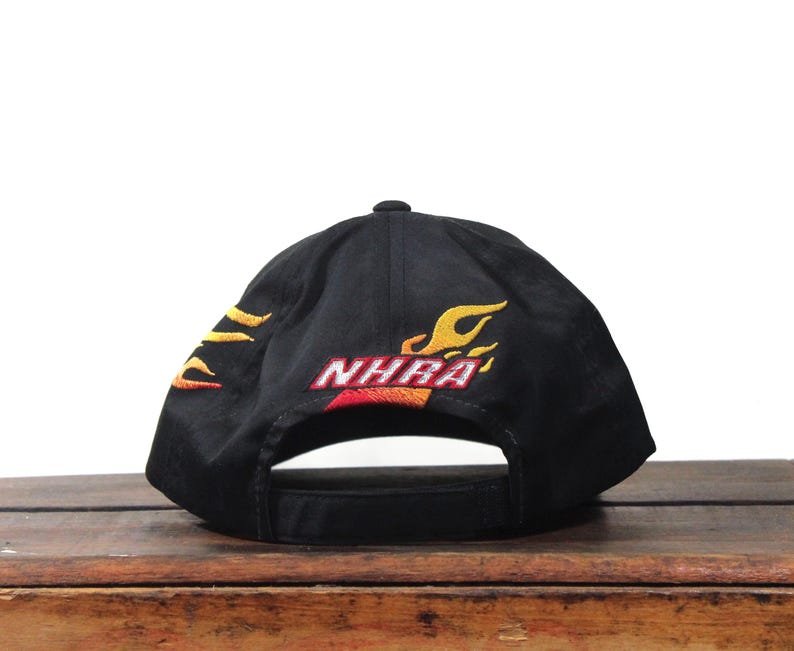 May include: Black baseball cap with embroidered flames in yellow and orange. The cap features the text "NHRA" in red and orange. The cap is sitting on a wooden surface.
