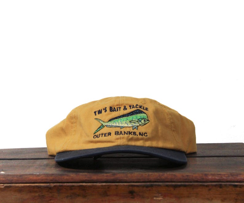 K&ouml;nnte beinhalten: Senfgelbe Kappe mit marineblauem Schirm. Die Kappe zeigt eine gestickte Fischgrafik in Gr&uuml;n und Blau mit dem Text "TW'S BAIT & TACKLE OUTER BANKS, NC" in Wei&szlig;.