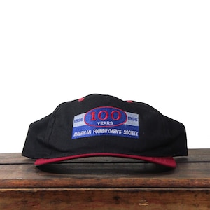 Puede incluir: Gorra de béisbol negra con visera y detalles en rojo. La gorra presenta un diseño bordado con el texto "100 YEARS" en rojo y "AMERICAN FOUNDRYMEN'S SOCIETY" en azul y blanco.