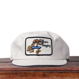 Op de afbeelding: Crèmekleurige baseballcap met een rechthoekige patch met een cartoonbever in werkkleding. De patch heeft de tekst "Billham Logging" in zwarte letters. De pet staat op een houten oppervlak.