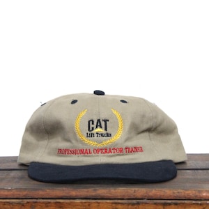 May include: A tan and black baseball cap with a black brim. The front of the cap features an embroidered yellow logo with the text "CAT Lift Trucks" and a wreath. The text "PROFESSIONAL OPERATOR TRAINER" is embroidered in red below the logo.