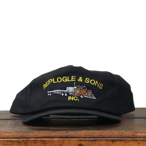 May include: Black cap with the embroidered text "REPLOGLE & SONS INC." and a graphic of a brown and orange semi-truck. The cap has a curved brim and a black rope detail along the edge.