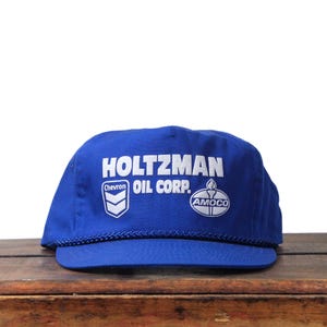 Puede incluir: Gorra de béisbol azul con el texto "HOLTZMAN OIL CORP." en blanco. La gorra presenta los logotipos de Chevron y Amoco. La visera es azul y la gorra tiene un detalle de cuerda alrededor de la base.