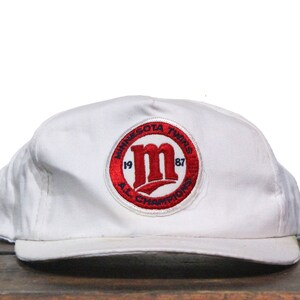 Puede incluir: Gorra de béisbol blanca con un parche rojo y azul de los Minnesota Twins 1987 AL Champions.