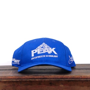 May include: A bright blue baseball cap featuring the white embroidered PEAK Antifreeze & Coolant logo with a mountain graphic on the front. The side panels display white embroidered text for BlueDEF Exhaust Fluid and a signature logo.