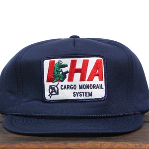 Puede incluir: Una gorra de camionero azul marino con un parche blanco que muestra un cocodrilo verde y el texto "LHA CARGO MONORAIL SYSTEM".