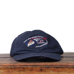 May include: A navy blue baseball cap with embroidered logos. The cap features the "Labatt Blue" logo and "USA Adult Hockey" text. The cap is sitting on a wooden surface.