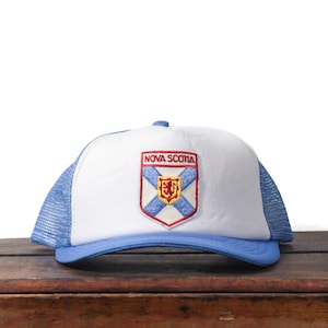 Peut inclure: Casquette de camionneur blanche et bleue avec une visière incurvée. Le chapeau présente un écusson avec les mots "Nova Scotia" et un écusson avec une croix bleue et un lion d'or.