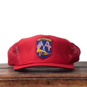 May include: A red trucker hat with a mesh back and a patch that reads "Gettysburg." The patch features an American flag, a Confederate flag, and a golden bugle on a blue shield.