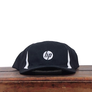 May include: A black baseball cap featuring the white HP logo embroidered on the front centre. The cap includes two vertical white accent stripes on the sides and a structured brim. The design is simple and functional with a classic six-panel construction.