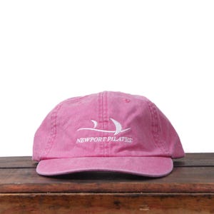 May include: A pink baseball cap with a curved brim. The cap features the white text "NEWPORT PILATES" and a stylized white graphic. The cap is made of a soft, textured material and sits on a wooden surface.