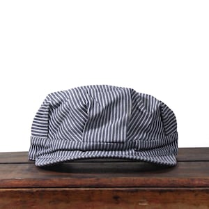 May include: A blue and white striped cap, reminiscent of a train conductor's hat, sits on a wooden surface. The cap features vertical stripes and a rounded crown, offering a classic, vintage aesthetic. The hat is made of fabric.