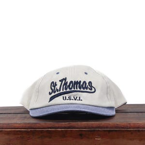 May include: A beige baseball cap with a blue brim and blue embroidered text on the front that reads St. Thomas and U.S.V.I. The cap features small blue ventilation eyelets on the crown and a structured six-panel design with a curved visor.