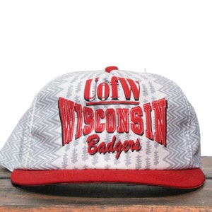 Puede incluir: Gorra de béisbol blanca y roja con un patrón de chevron gris. La gorra tiene el texto "U of W WISCONSIN Badgers" en rojo.