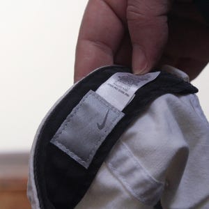 Puede incluir: Gorra de b&eacute;isbol blanca con visera negra. Se ve una etiqueta gris con el logotipo de Nike. La gorra est&aacute; sujeta por una mano.