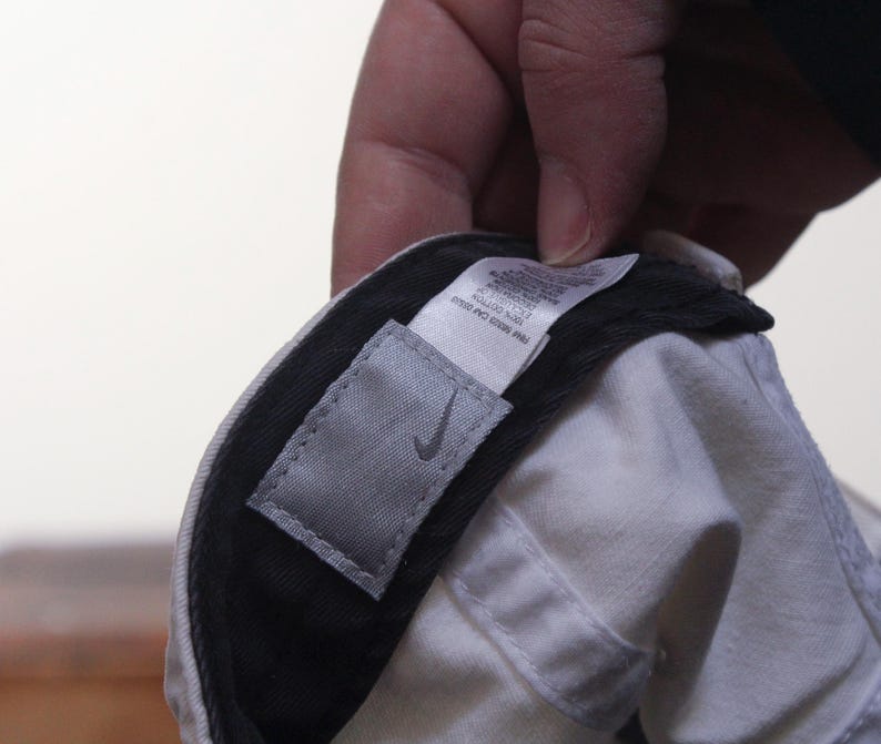 Puede incluir: Gorra de b&eacute;isbol blanca con visera negra. Se ve una etiqueta gris con el logotipo de Nike. La gorra est&aacute; sujeta por una mano.