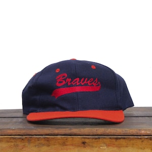 以下が含まれることがあります： 紺色のベースボールキャップに赤いつばと赤い刺繍の「ブレーブス」ロゴが付いている