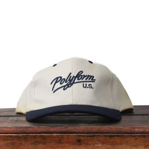 Puede incluir: Gorra de béisbol color crema con visera azul marino. La gorra presenta la palabra "Polyform" en escritura azul marino, con "U.S." debajo. El sombrero está sobre una superficie de madera.