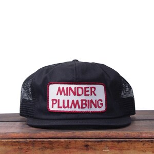 Może przedstawiać: Czarna czapka typu trucker z siatkowym tyłem i prostokątną naszywką. Na naszywce widnieje napis "MINDER PLUMBING" w kolorze czerwonym na białym tle.