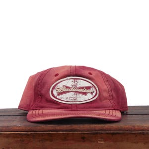 May include: A faded red baseball cap featuring a white oval patch on the front. The patch contains the red Budweiser logo with a distressed, weathered texture. The cap has a curved brim and stitched panels, displaying a worn, vintage appearance.