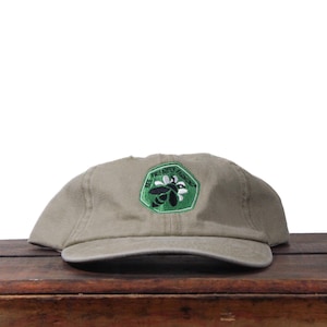 Puede incluir: Gorra de béisbol beige con visera curva. Cuenta con un parche verde con una abeja y las palabras "BEE FRIENDLY FARMING" en blanco. Se encuentra sobre una superficie de madera.