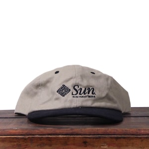 May include: A tan baseball cap with a black brim. The cap features the embroidered text "Sun microsystems" in black. The hat is sitting on a wooden surface against a white background.