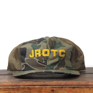 May include: Camouflage baseball cap with the text "JROTC" in yellow block letters. The cap features a mesh back and a curved brim. The front panel has a green, brown, and black camouflage pattern.