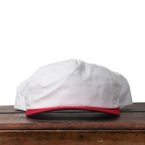 May include: A white baseball cap with a red brim sits on a wooden surface. The cap is made of a soft, slightly wrinkled fabric. The red brim provides a striking contrast to the white fabric.