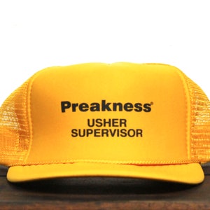 May include: A yellow trucker hat with the text "Preakness USHER SUPERVISOR" in black. The hat has a mesh back and a curved brim. The hat is likely a promotional item or souvenir.