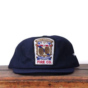May include: A navy blue baseball cap with a flat brim. The cap features a rectangular patch with the text "MILLSBORO DELAWARE FIRE CO." and a crest design with two wheels and a banner.