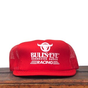 May include: A red trucker hat with a white bull's head logo and the text "BULL'S-EYE BARBECUE SAUCE RACING" in white. The hat has a mesh back and a flat brim. The hat is sitting on a wooden surface.