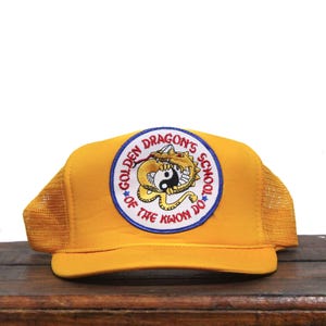 Pode incluir: Um boné de caminhoneiro de malha amarela com um remendo que diz "Golden Dragon's School of the Kwoon Do" e apresenta um dragão amarelo segurando um símbolo yin yang.