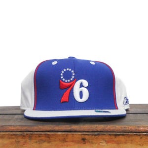 Könnte beinhalten: Eine blaue und weiße Baseballkappe mit einem roten und weißen "76" Logo und einem Sternenkranz. Die Kappe ist im Fitted-Stil.