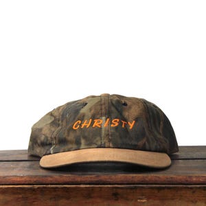 May include: A camouflage baseball cap with the name "CHRISTY" embroidered in orange thread. The cap has a brown suede peak and a classic six-panel design. The hat is set against a white background.
