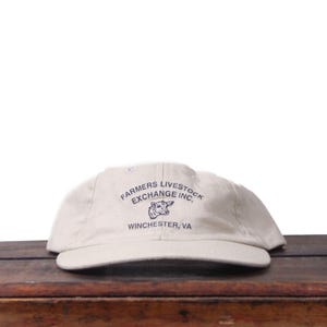 May include: A beige baseball cap with a curved brim. The cap features the text "FARMERS LIVESTOCK EXCHANGE INC." in navy blue, along with a graphic of a cow and the location "WINCHESTER, VA."