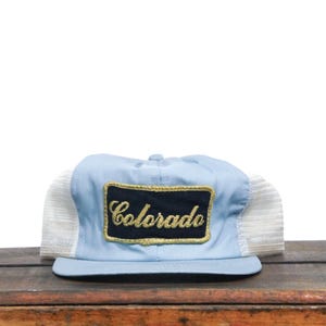 Puede incluir: Una gorra de camionero azul claro con una malla blanca en la parte posterior. La gorra tiene un parche bordado en negro y dorado que dice "Colorado" en la parte delantera.