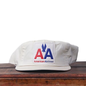 May include: Cream-colored baseball cap featuring the American Airlines logo. The logo is a square patch with the letters "AA" in red and blue, and a stylized eagle. The text "American Airlines" is below the logo.