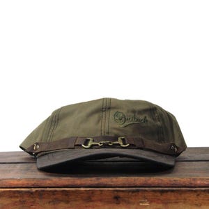 May include: Olive green newsboy cap with a dark brown leather band and a metal buckle detail. The cap has the word "Outback" embroidered in green on the side. The cap is sitting on a wooden surface.