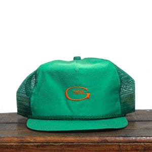 Puede incluir: Una gorra de camionero de malla verde con un logotipo bordado en naranja que dice "PAPER G".