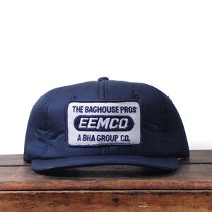 May include: A navy blue baseball cap with a rectangular patch. The patch has the text "THE BAGHOUSE PROS" above the word "EEMCO" in white and blue. Below the word "EEMCO" is the text "A BHA GROUP CO."