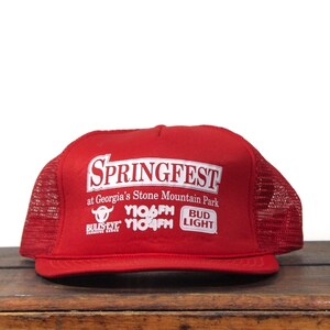 May include: A red trucker hat with a white graphic that reads "SPRINGFEST at Georgia's Stone Mountain Park". The hat also features the logos of Y106FM, Bulis Eye Barbecue, and Bud Light. The hat has a mesh back.