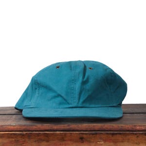 May include: Teal baseball cap with a curved peak. The cap is made of a solid-coloured fabric and has two small ventilation holes on top. It is displayed on a wooden surface.