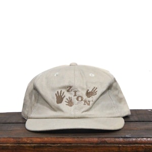 May include: A beige baseball cap with brown embroidery of the word "Zion" and handprints.