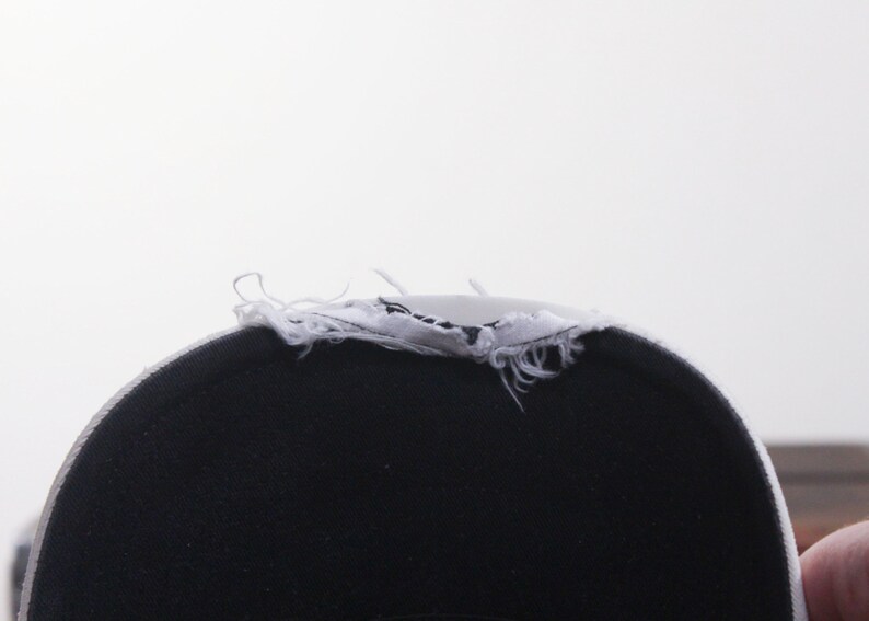 Puede incluir: Primer plano de una gorra de b&eacute;isbol negra con un borde deshilachado blanco. El interior de la gorra es negro y el borde deshilachado es blanco con costuras visibles. El fondo es blanco liso.