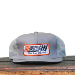 Puede incluir: Gorra de camionero gris con parte trasera de malla y visera plana. La parte delantera de la gorra presenta un parche rectangular con la palabra "ECHO" en blanco y negro, y el texto "First, from the start" en naranja.